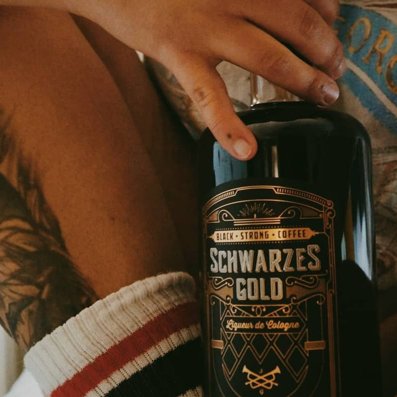 Schwarzes Gold Espresso-Likör Flasche, gehalten von tätowierter Hand, mit Fokus auf Bein in gestreiften Socken, Kaffeelikör aus Bio-Fairtrade-Kaffee, ideal für Cocktails und puren Genuss.